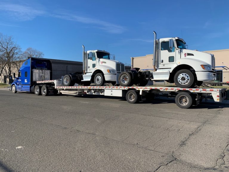 Two white Kenworth semi trucks shipped on a step deck trailer.